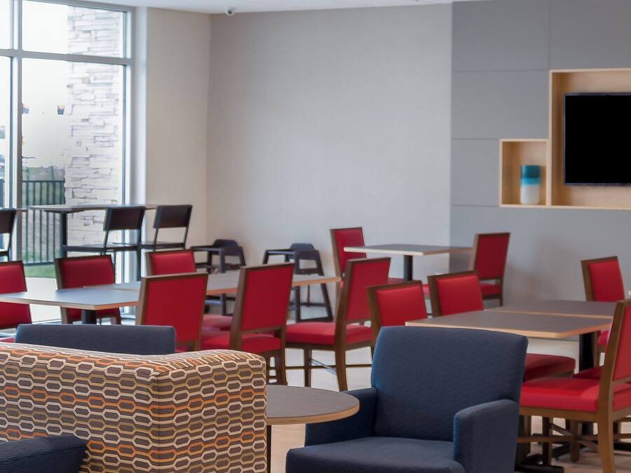 A modern dining area with red chairs and tables, a large flat-screen TV mounted on the wall, and a patterned ottoman in the foreground.