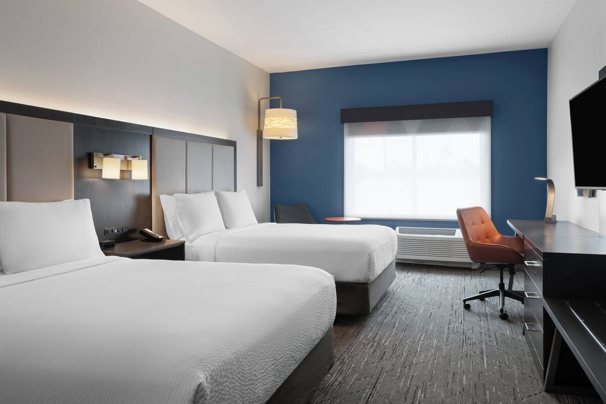 A hotel room with two beds, a desk, a chair, a TV, and a large window.