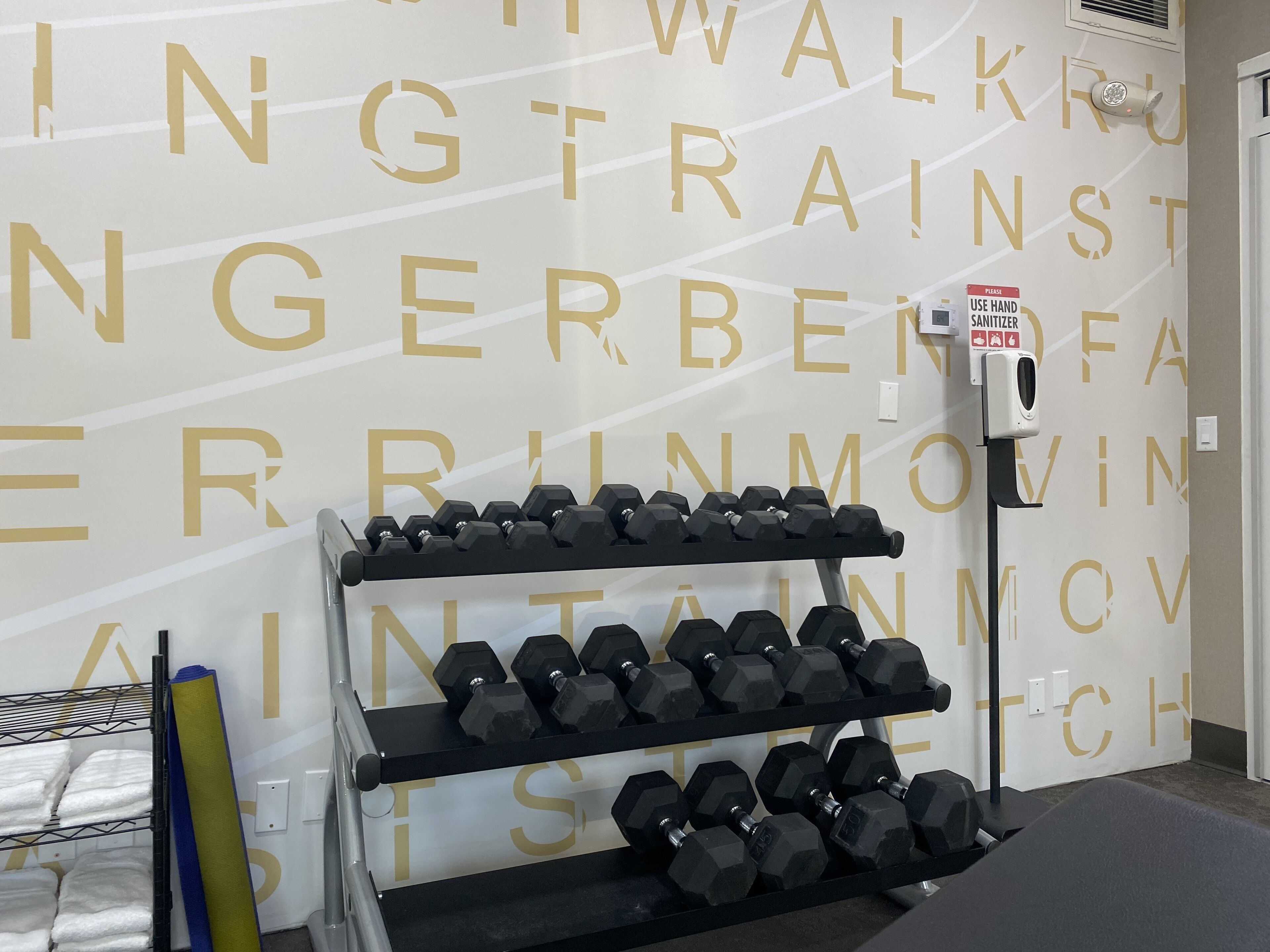 Uma academia com uma parede exibindo letras douradas grandes, um suporte de halteres e um dispenser de álcool em gel.