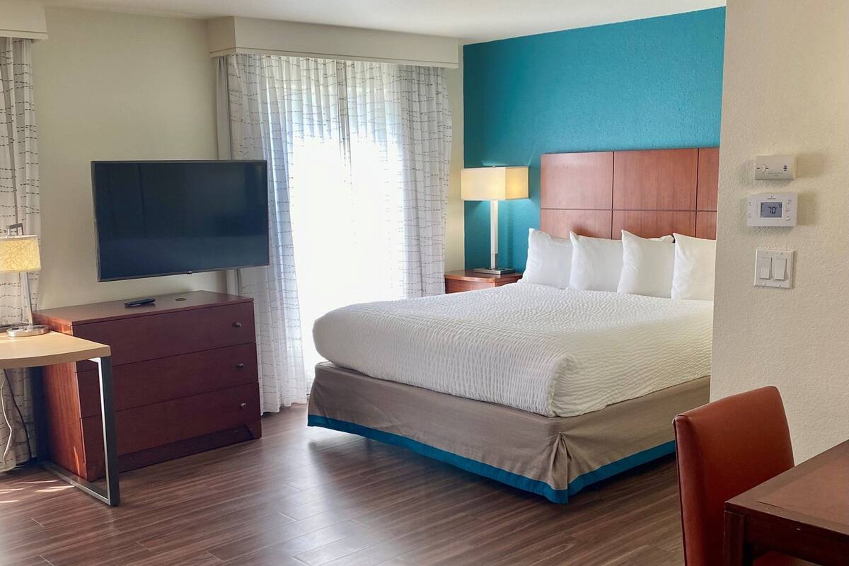 Ein Hotelzimmer mit einem Bett, einem Fernseher auf einer Holzkommode, einem Schreibtisch, einer Lampe und einem Fenster mit Vorhängen.
