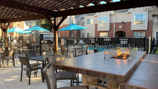 Une terrasse extérieure avec une table en pierre, des chaises et un foyer. On aperçoit des parasols et une piscine à l’arrière-plan.