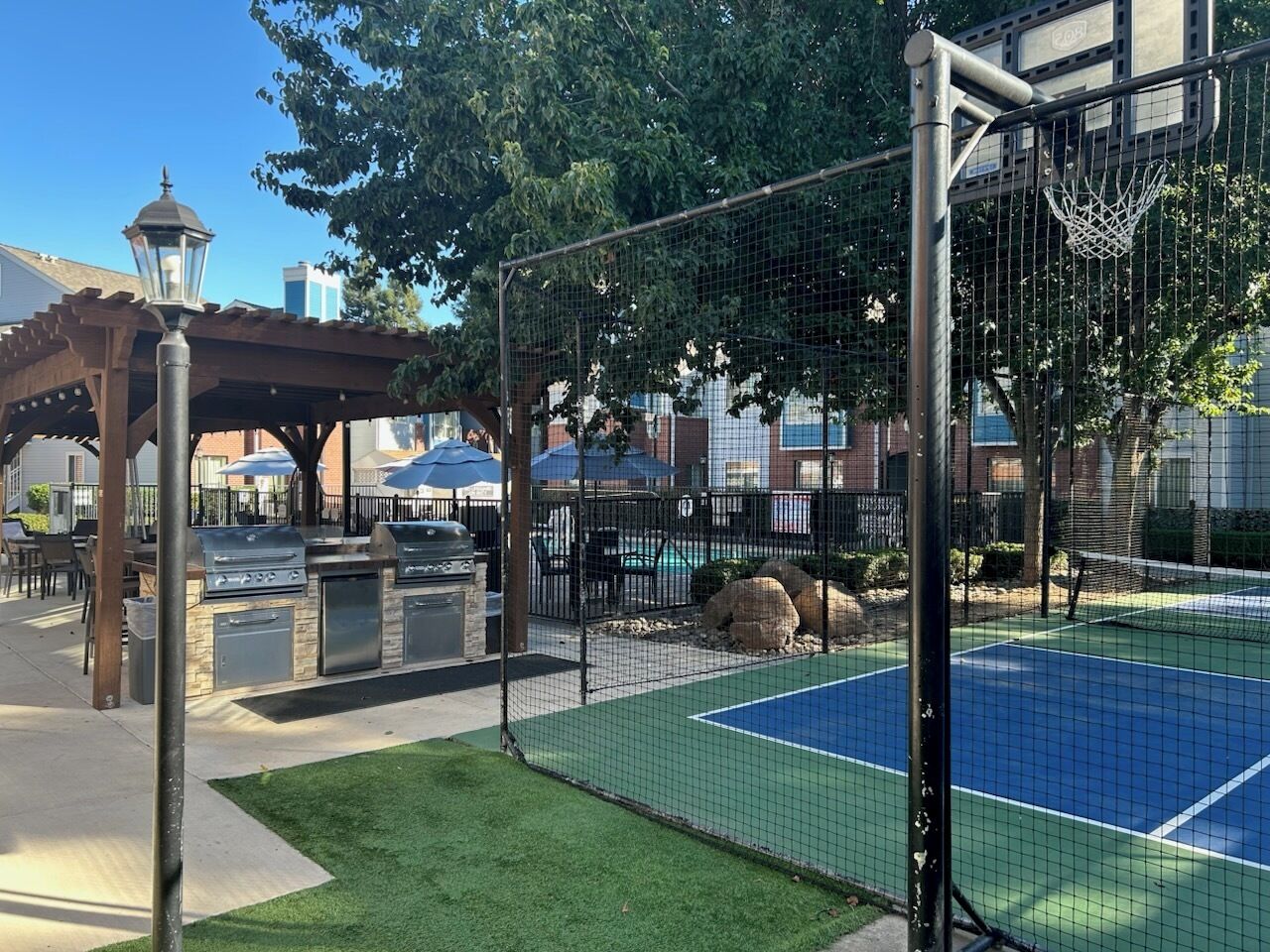 Een buitenbasketbalveld met een net, een barbecueplaats en een pergola met parasols.