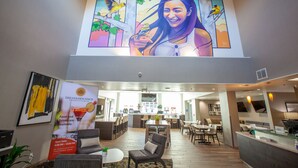 A modern hotel lobby with a large mural, a reception desk, and a dining area.