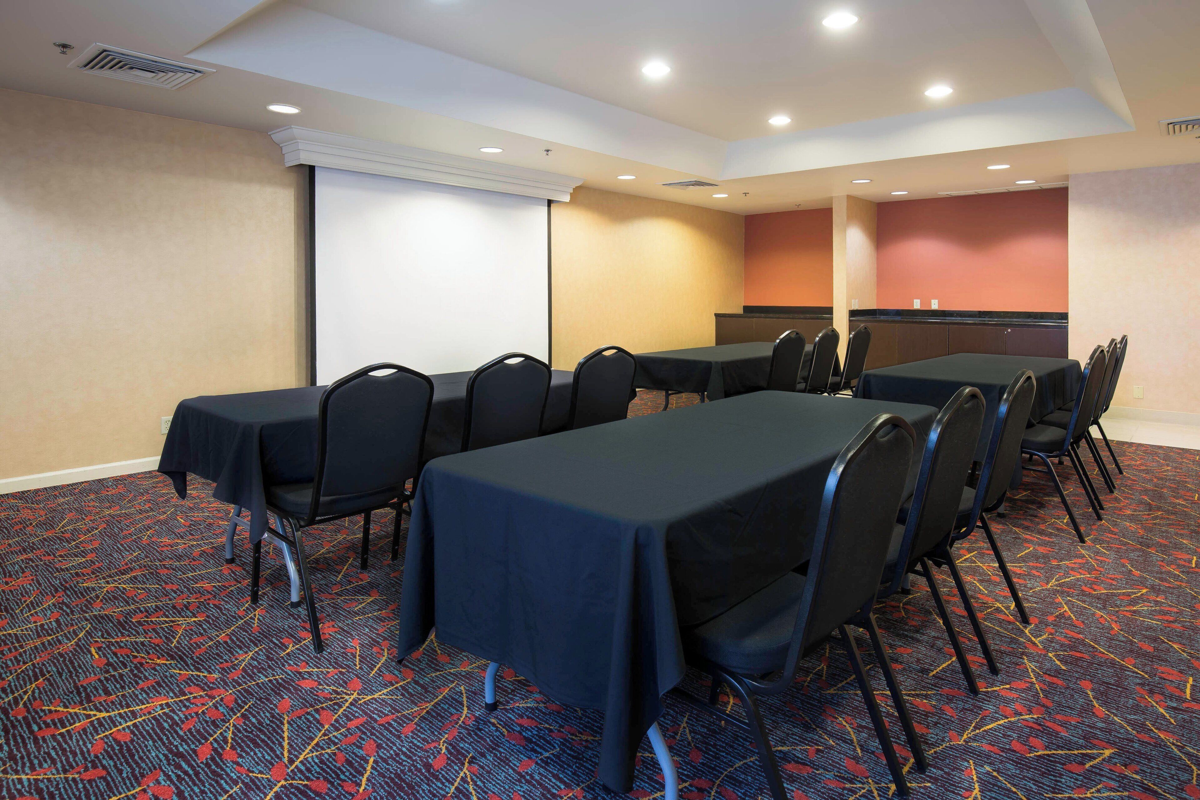 Un salón de conferencias con mesas y sillas, piso alfombrado y una pantalla para proyector.