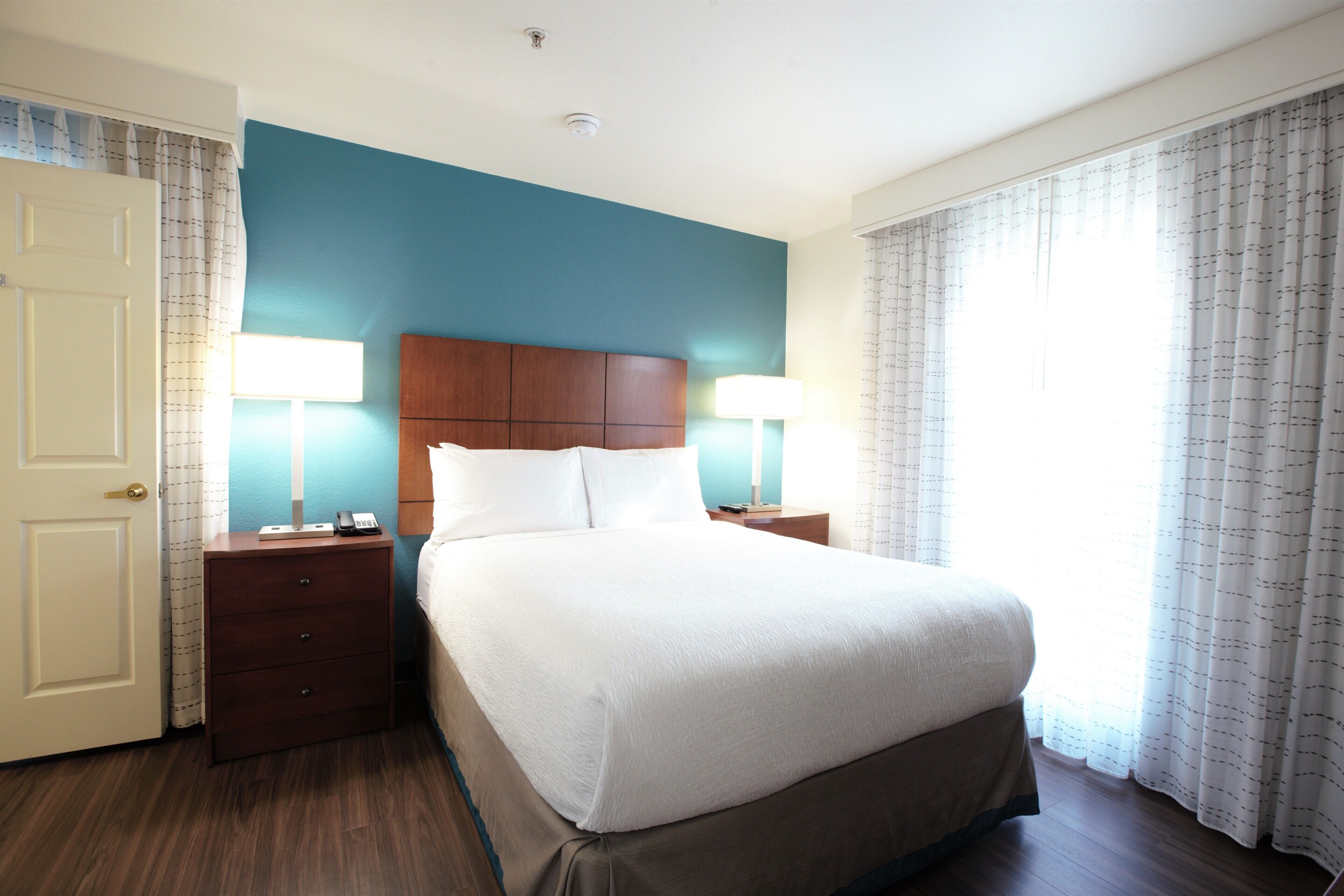 Une chambre d’hôtel avec un grand lit, deux tables de chevet avec des lampes, une tête de lit en bois et une vue sur l’extérieur par la fenêtre.