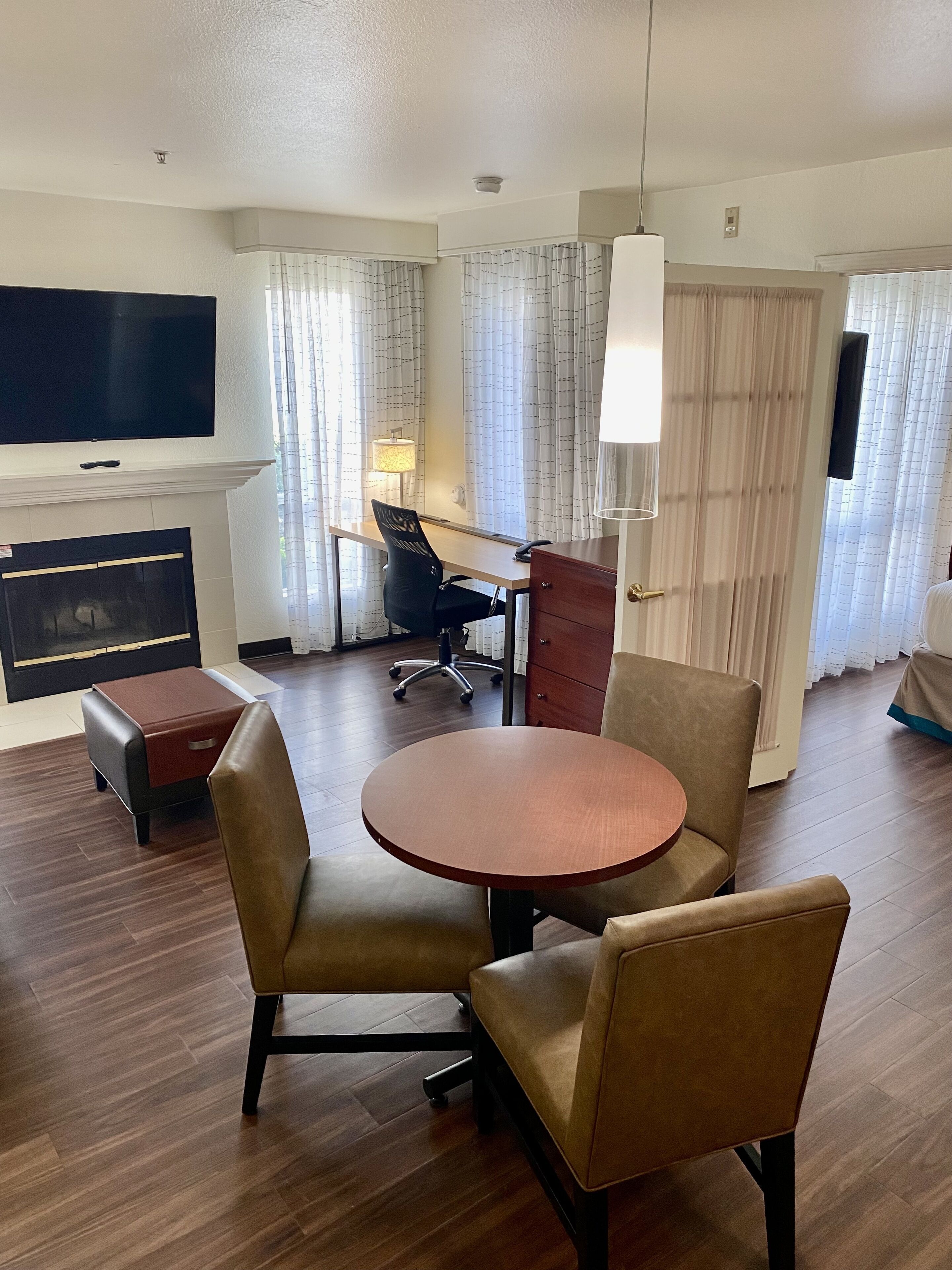 Une chambre d’hôtel dotée d’un coin repas comprenant une table ronde en bois et quatre chaises, un bureau avec une chaise, un téléviseur à écran plat fixé au mur, une cheminée et une grande fenêtre avec des rideaux.