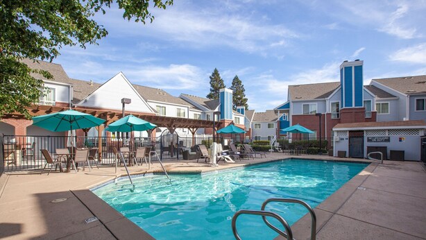 A swimming pool with lounge chairs and umbrellas, surrounded by buildings with a brick facade and blue accents.