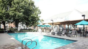 A swimming pool with lounge chairs and umbrellas, surrounded by trees and a building in the background.