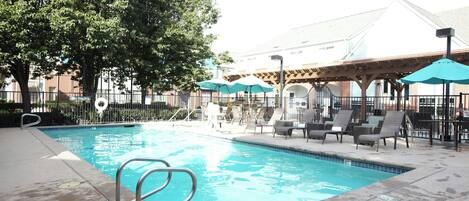 A swimming pool with lounge chairs and umbrellas, surrounded by trees and a building in the background.