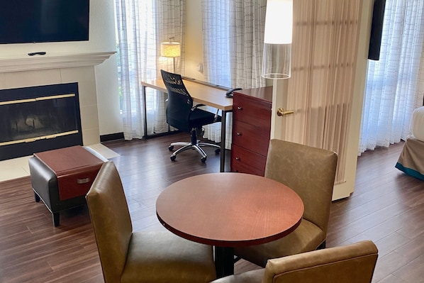 Une chambre d’hôtel avec une table ronde en bois, deux chaises beiges, un bureau avec une chaise, un téléviseur fixé au mur, une cheminée et une fenêtre avec des rideaux.