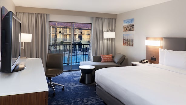 A hotel room with a large bed, a desk with a computer, a chair, a small round table, and a balcony with a pool view.