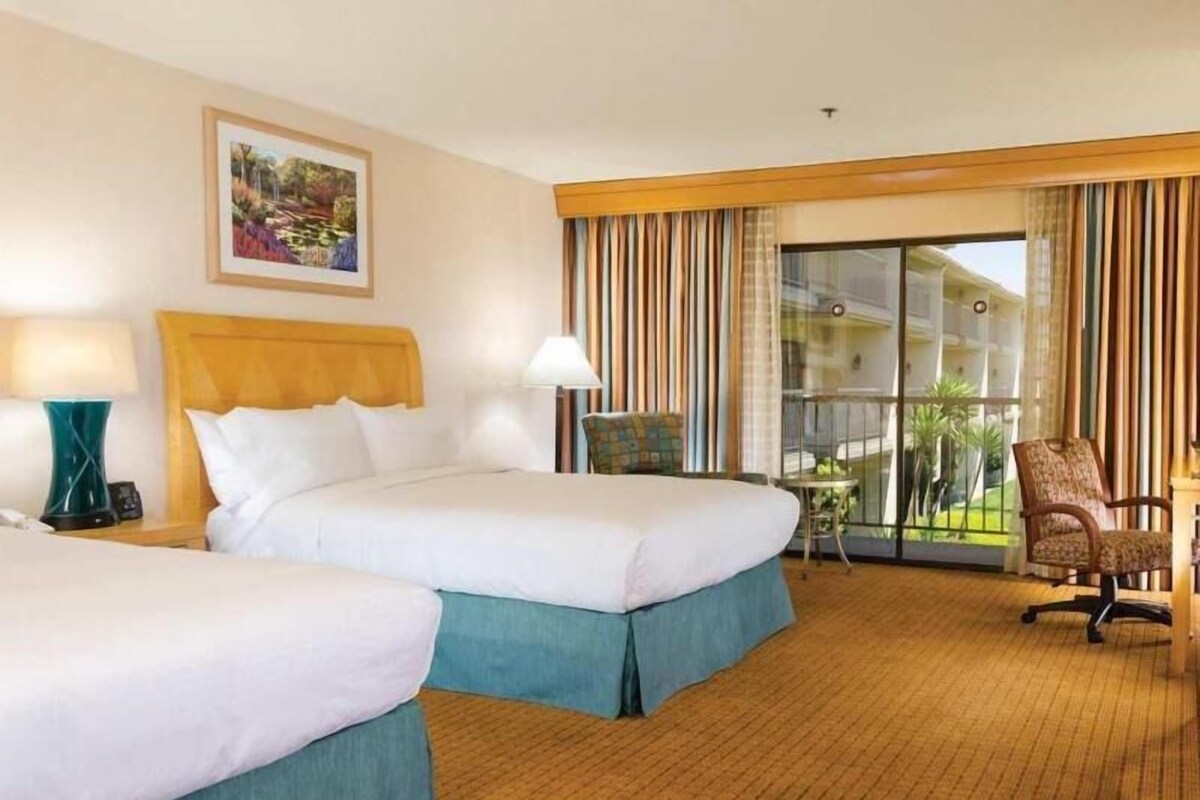 A hotel room with two beds, a small table, a chair, a painting on the wall, and a view through the window.
