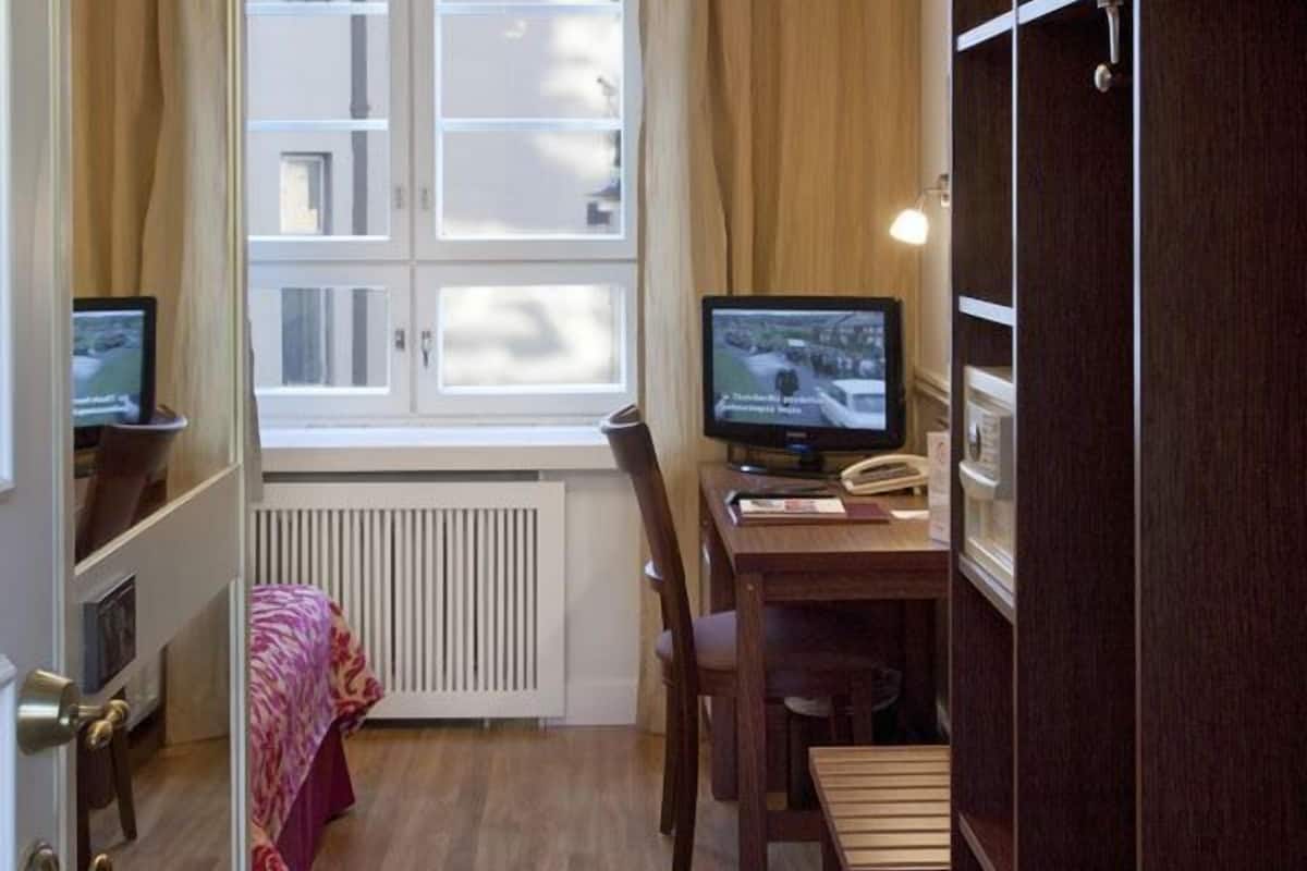 A compact room with a wooden floor, a desk with a computer, a chair, a bed with a red blanket, a window with curtains, and a wardrobe.