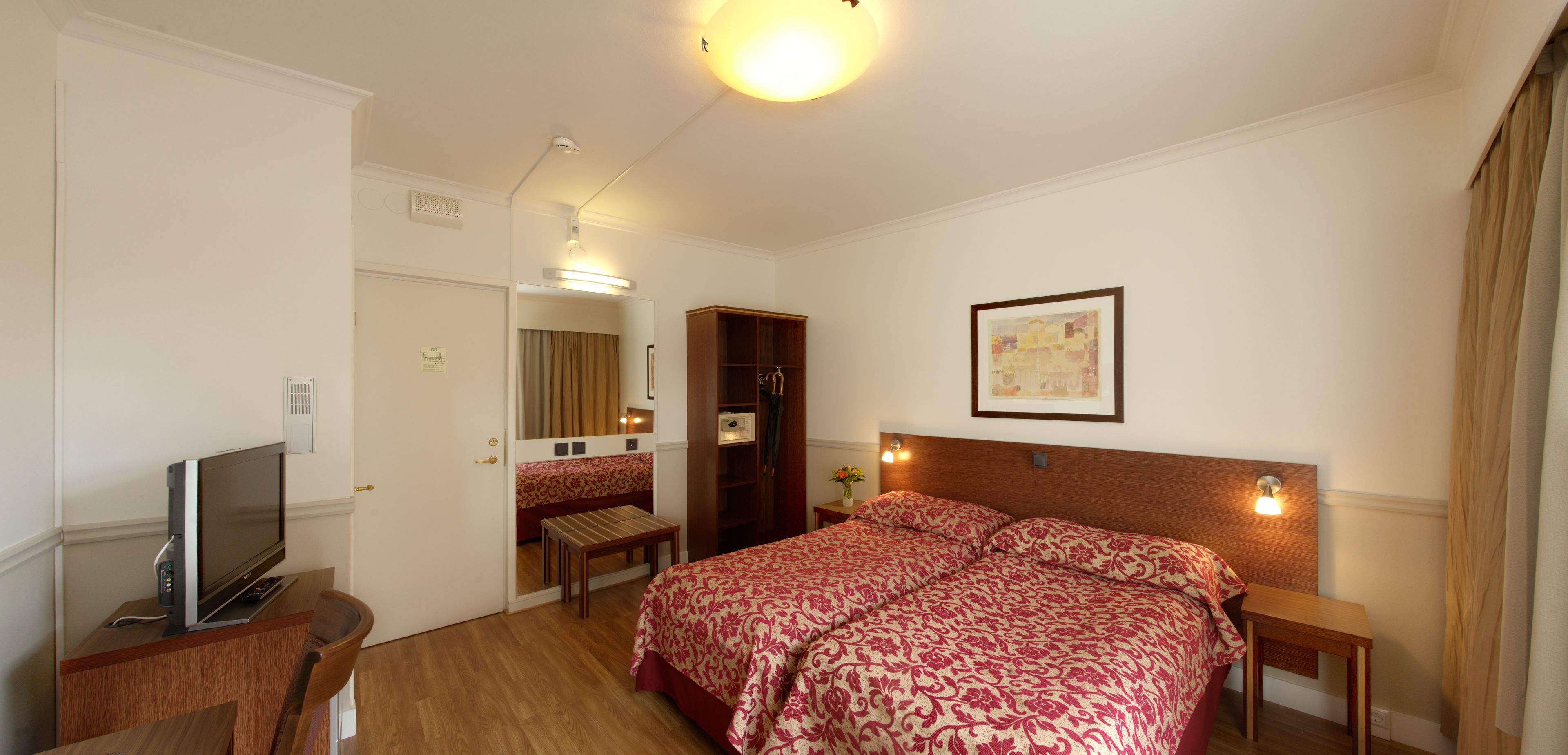 Una habitación de hotel con cama, mesitas de noche, televisión, escritorio y espejo.
