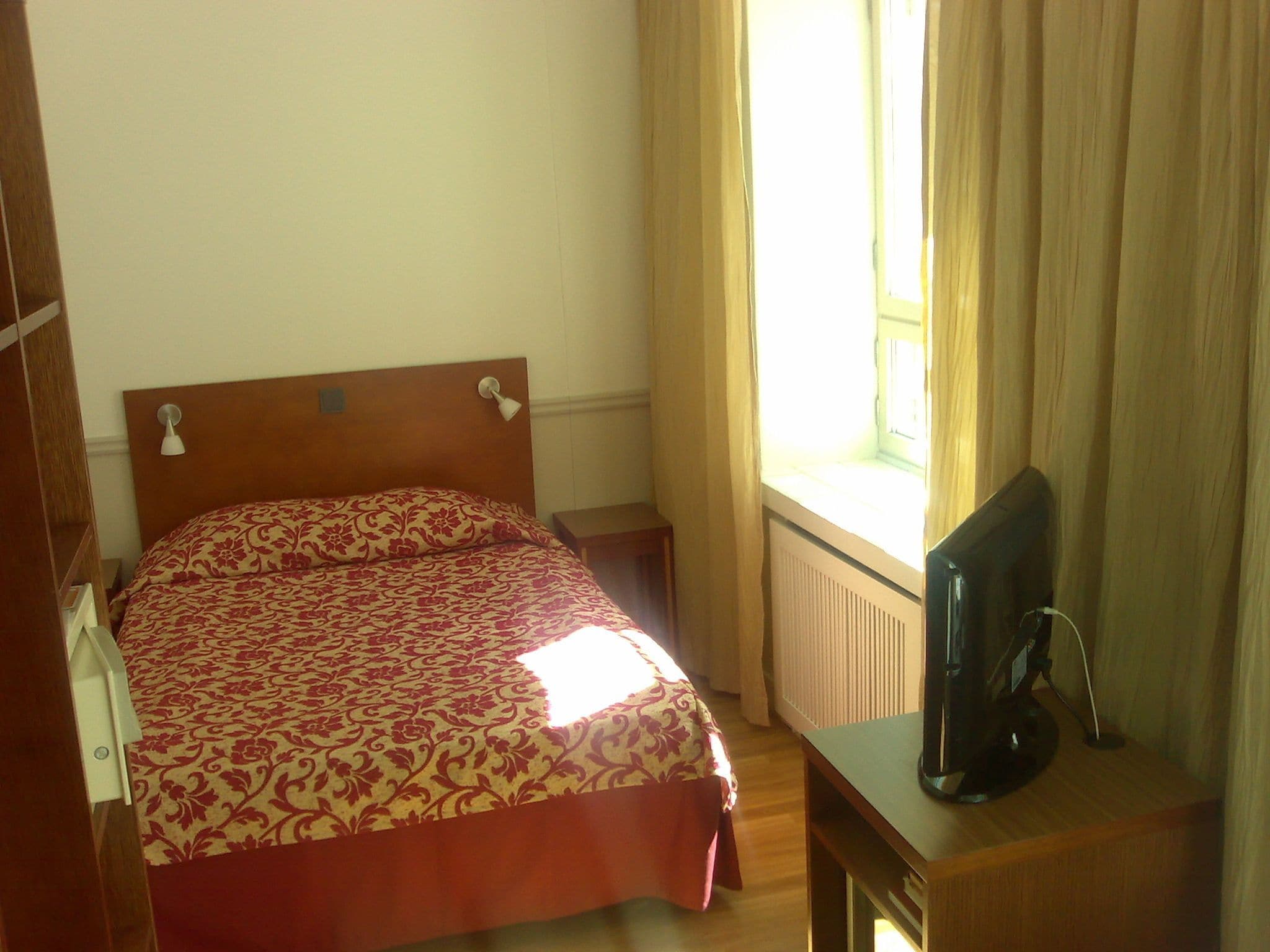 A bedroom with a bed, a bedside table, a TV on a stand, and a window with curtains.