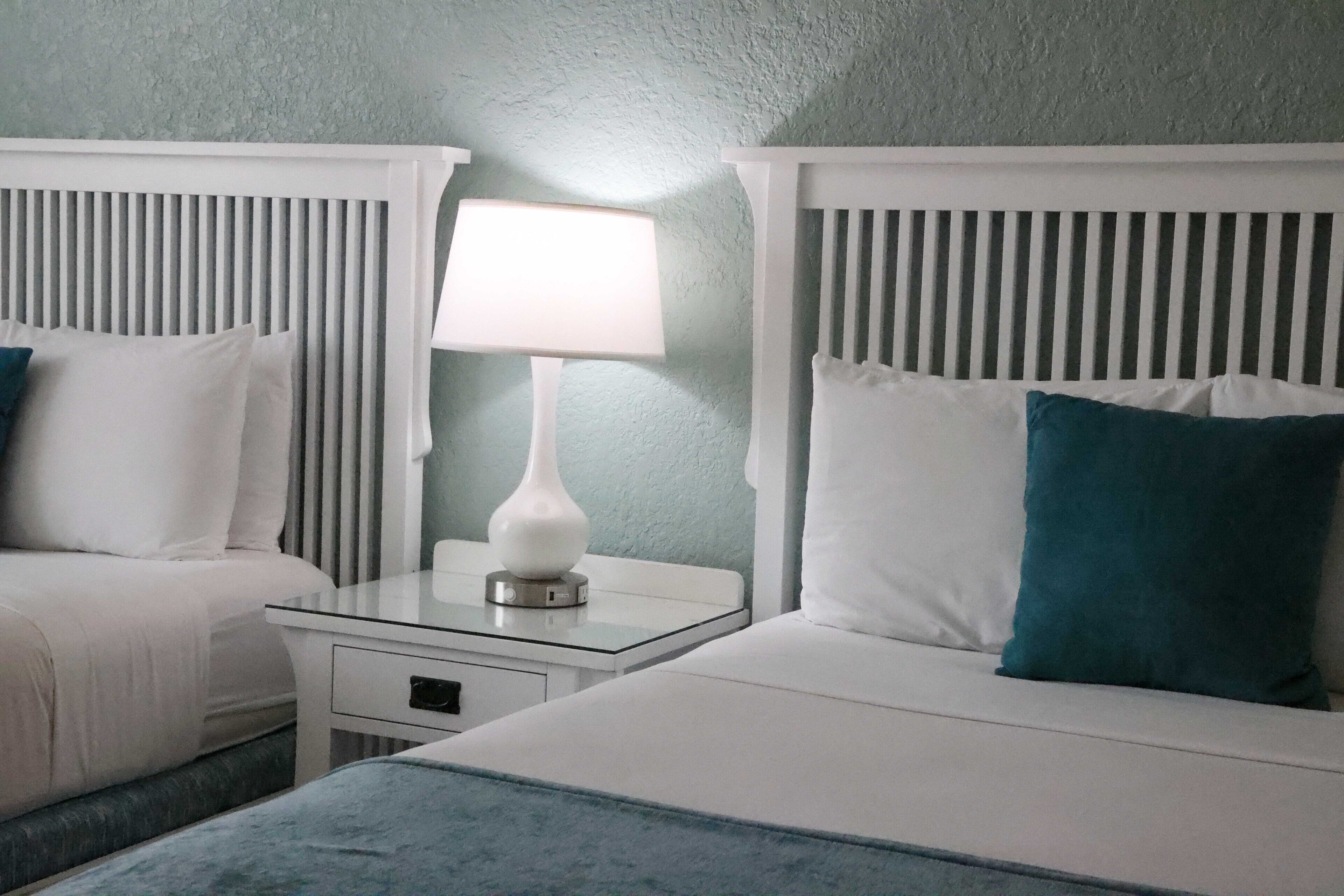 A hotel room with two beds, each with white bedding and teal pillows. There is a nightstand with a white lamp between the beds.