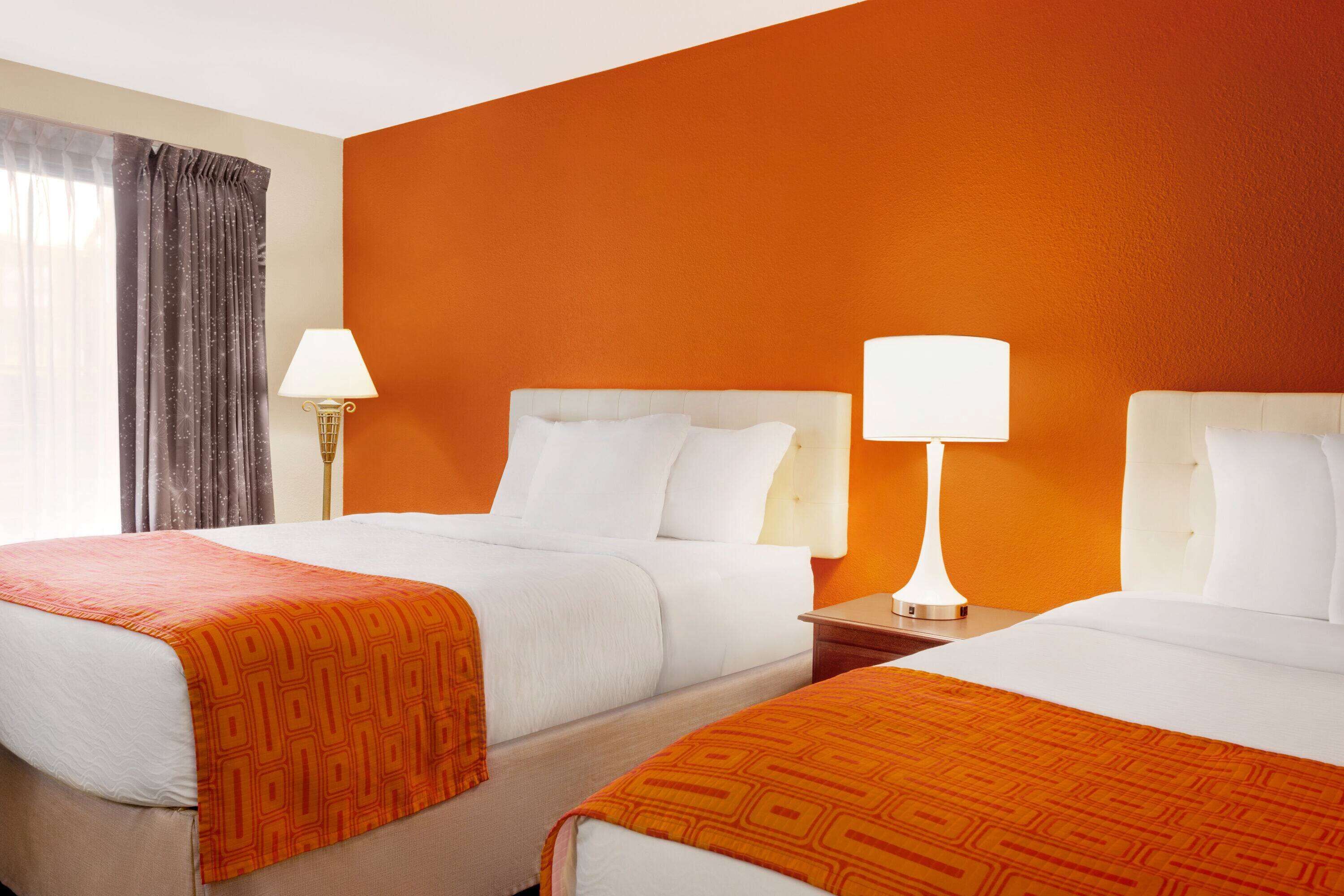 A hotel room with two beds, each with white bedding and orange throw blankets. There are two bedside tables with lamps, and a window with curtains.