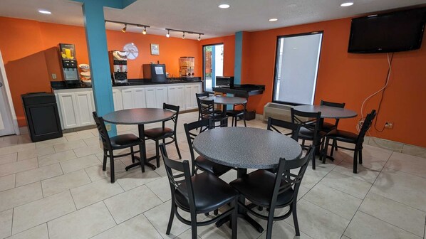 Un comedor con mesas y sillas redondas, una zona de cocina con una máquina de café, y un televisor montado en la pared.