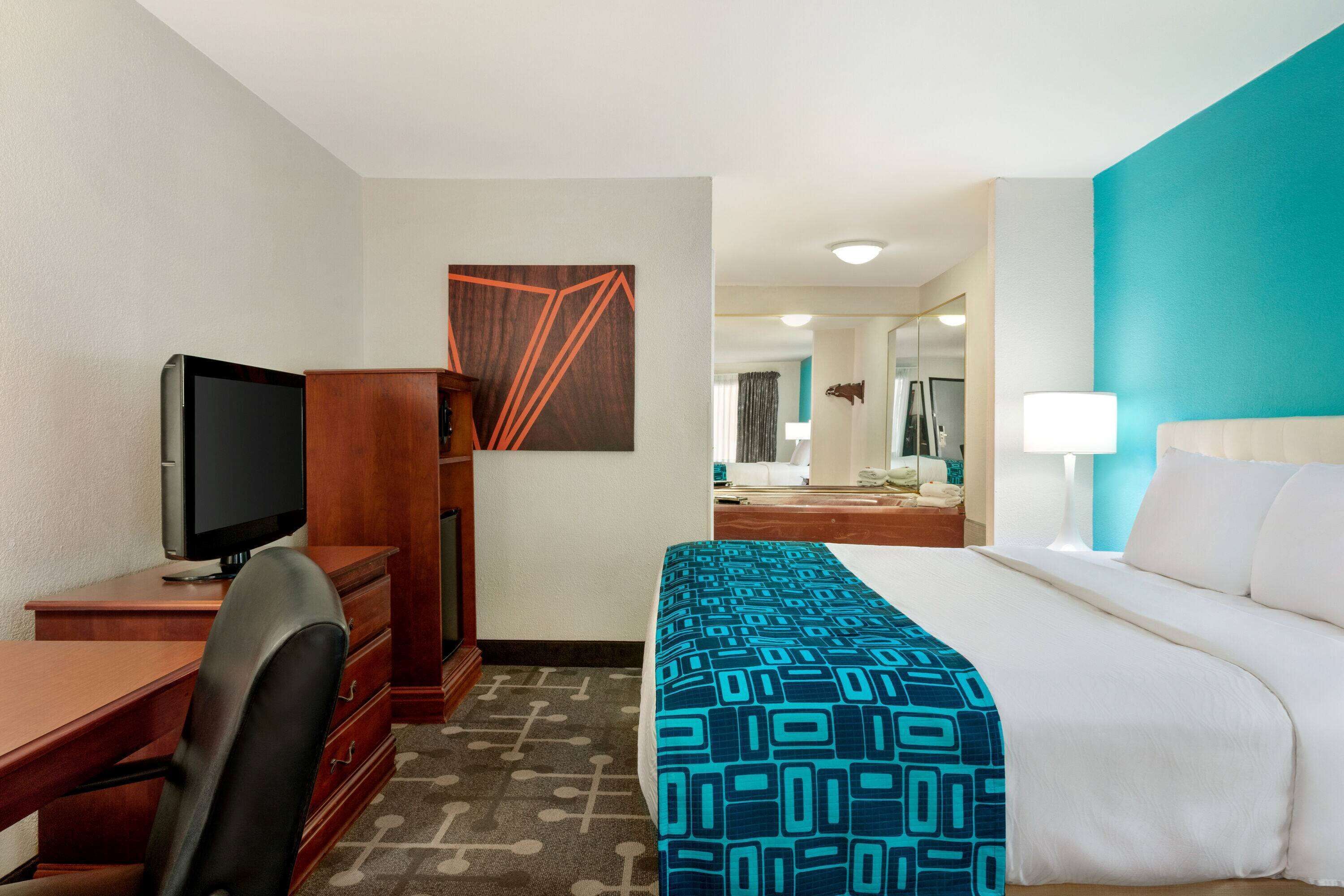Une chambre d’hôtel avec un lit, un bureau avec une télévision, une chaise, une commode et une salle de bain visible au fond.