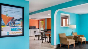 A hotel lobby with teal walls, a framed advertisement, a dining area with chairs and a table, and a kitchen in the background.