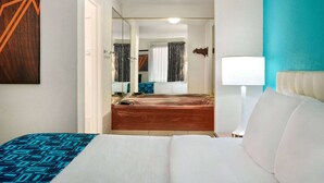 A hotel room with a bed, a mirror, a bathroom with a bathtub, and a nightstand with a lamp.