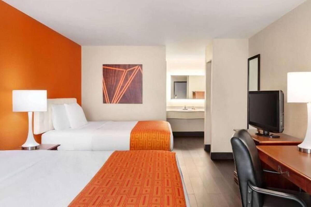 Habitación de hotel con dos camas, un escritorio con una silla, un televisor y un baño visible a través de una puerta abierta.