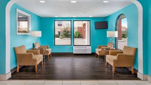 A lobby with teal walls, dark wooden flooring, and beige armchairs. There are two large windows with a view of the outside, a white air conditioning unit, and a flat-screen TV mounted on the wall.
