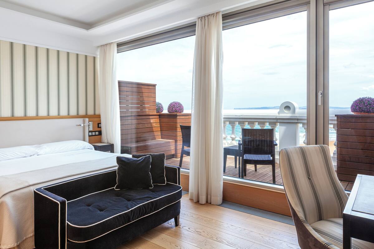 A hotel room with a bed, a black sofa, a striped armchair, and a balcony with a view of the sea.