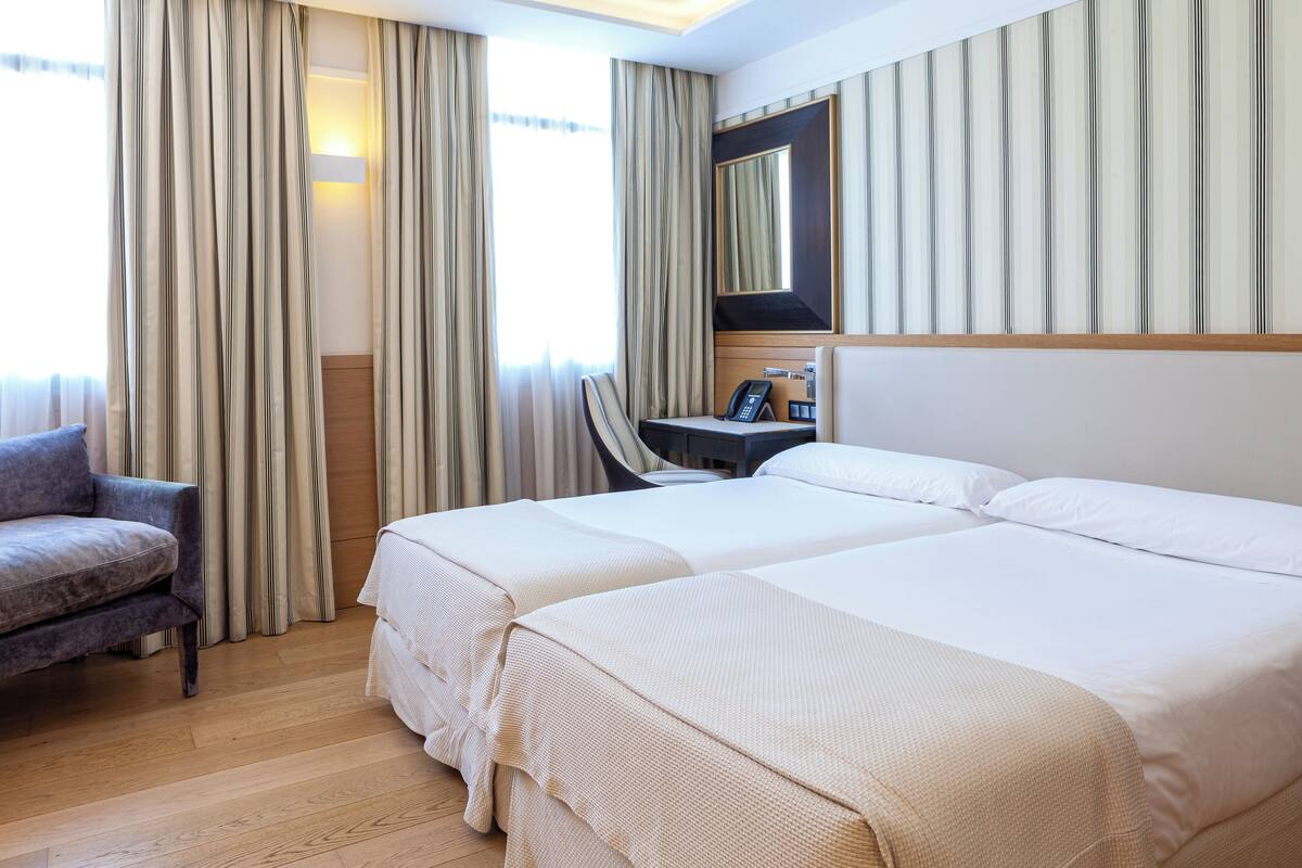 A hotel room with two beds, a desk with a phone, a chair, and a large window with curtains.