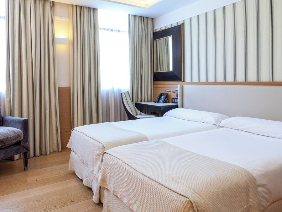 Een hotelkamer met twee bedden, een bureau met telefoon, een stoel en een groot raam met gordijnen.