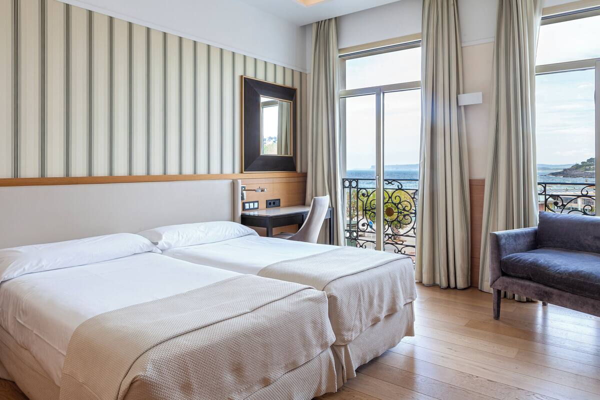 A hotel room with two beds, a desk, a chair, a large window with curtains, and a view of the sea.