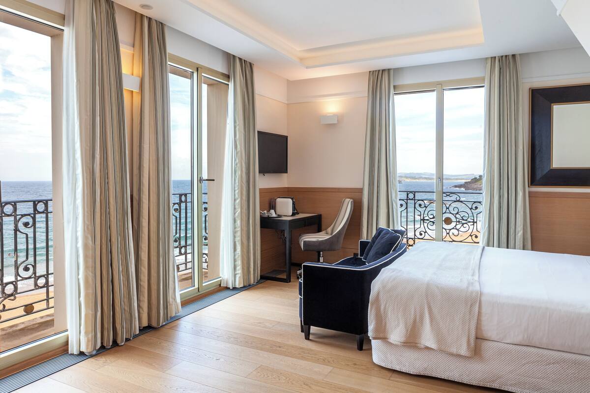 A hotel room with a bed, a desk, a chair, and a TV. The room has large windows with curtains and a view of the sea.