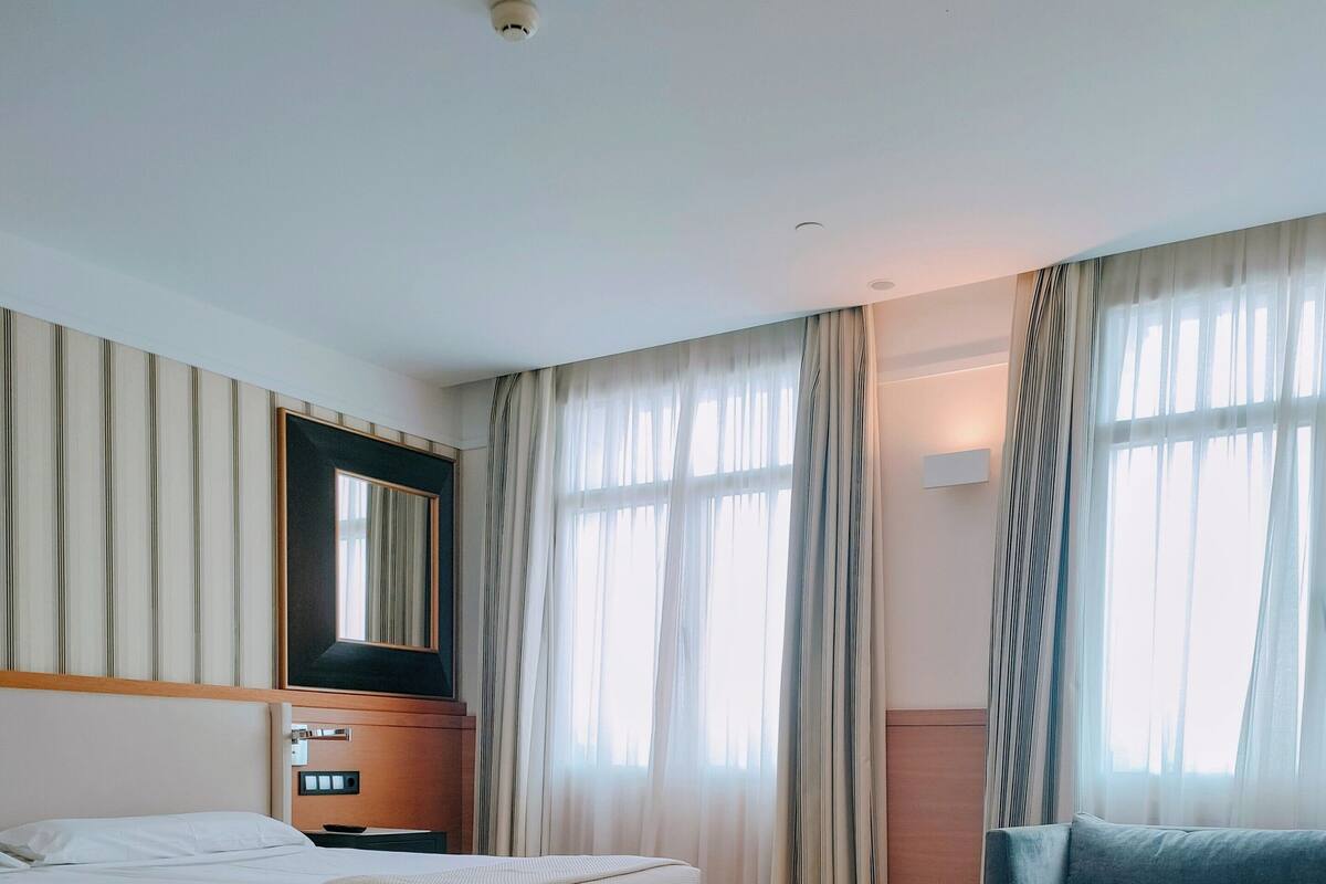 A hotel room with two beds, a wooden floor, a large window with curtains, a mirror, and a wall-mounted light.