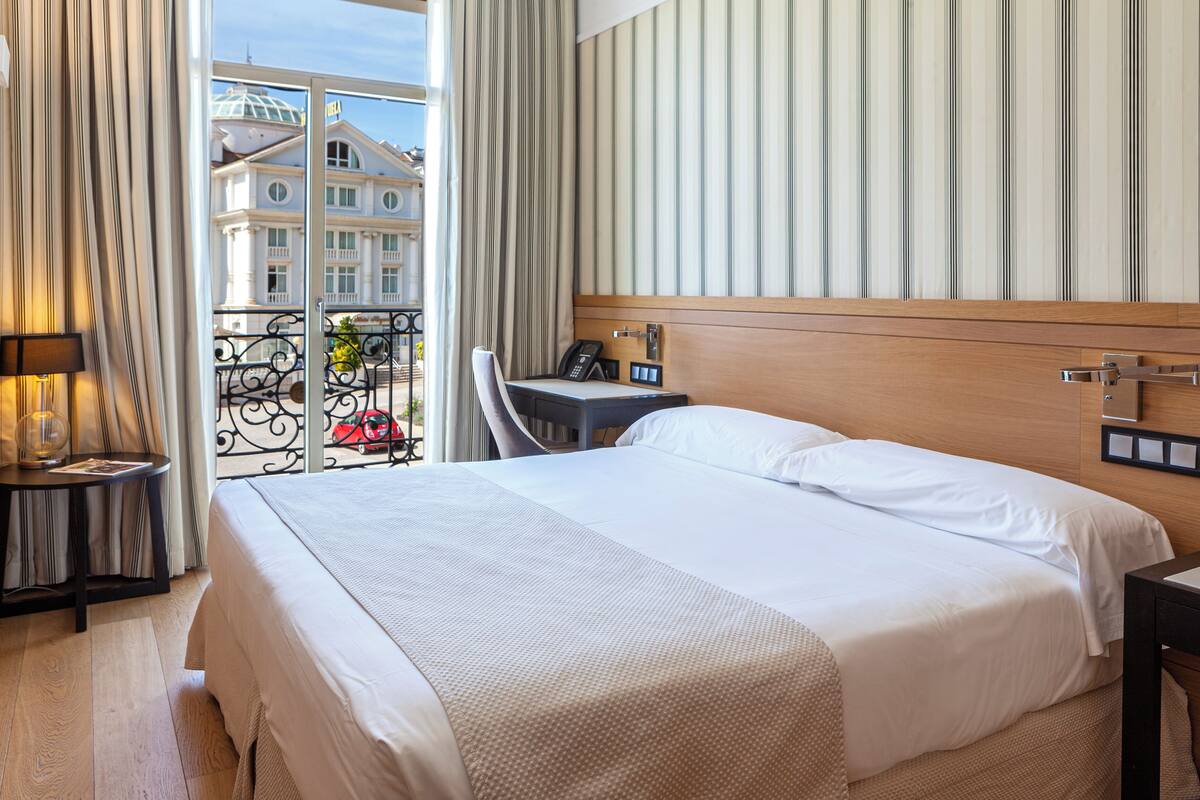 A hotel room with a large bed, a bedside table with a lamp, a chair, and a view of a building through a window.