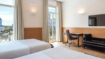 Quarto de hotel com duas camas, uma escrivaninha, uma cadeira, televisão e varanda com vista para a cidade.