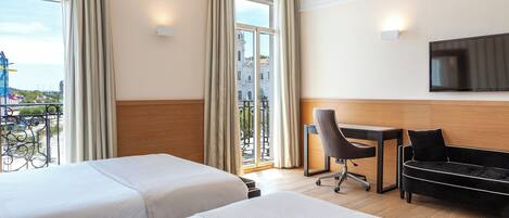 A hotel room with two beds, a desk, a chair, a TV, and a balcony with a view of the city.