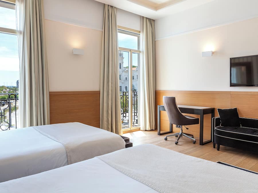 Quarto de hotel com duas camas, uma escrivaninha, uma cadeira, televisão e varanda com vista para a cidade.