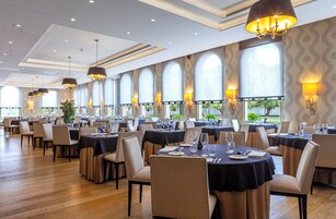 Un comedor amplio con varias mesas dispuestas para comer, ventanas grandes con arcos y candelabros modernos.