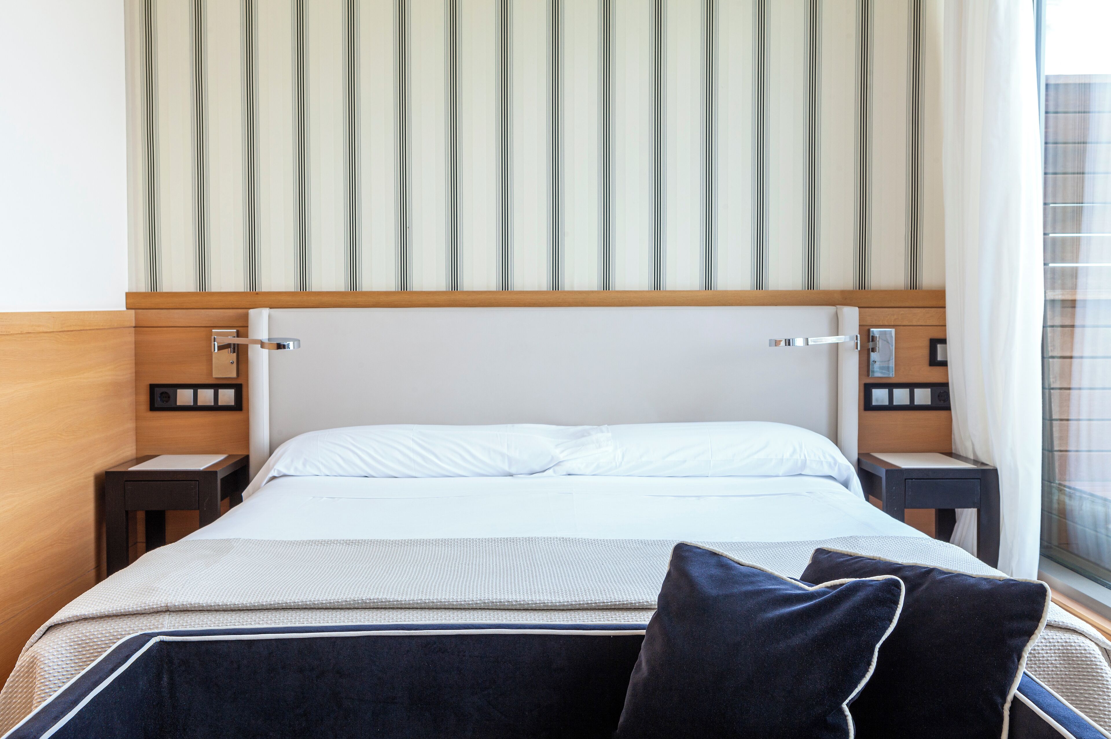 Habitación de hotel con una cama grande, cabecero de madera, dos mesitas de noche y un ventanal con cortinas.
