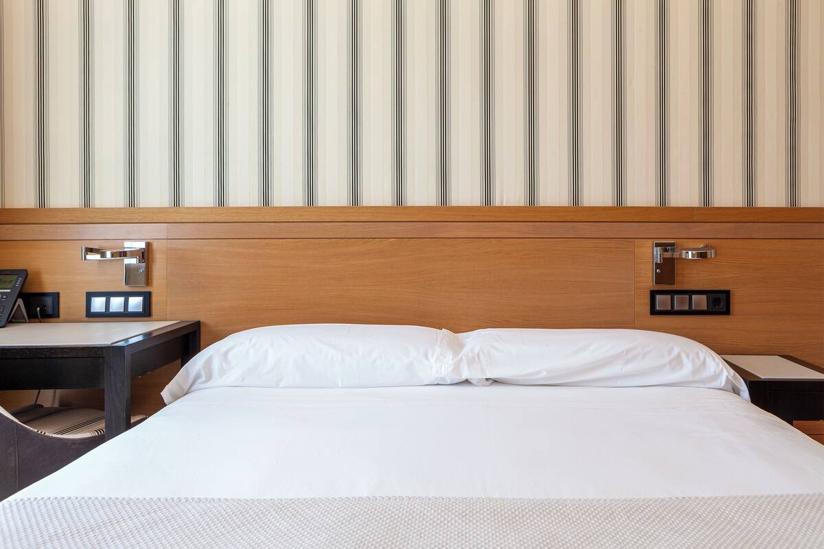 A hotel room with a wooden headboard, a bed with white linens, a nightstand with a phone and a lamp, and a wall-mounted light switch.