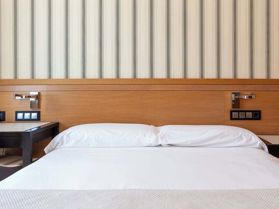 Una habitación de hotel con cabecera de madera, cama con ropa de cama blanca, mesita de noche con teléfono y lámpara, y un interruptor de luz en la pared.