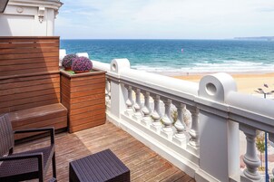 Un balcón con piso de madera, barandas blancas, mobiliario de mimbre y vista a la playa y al océano.