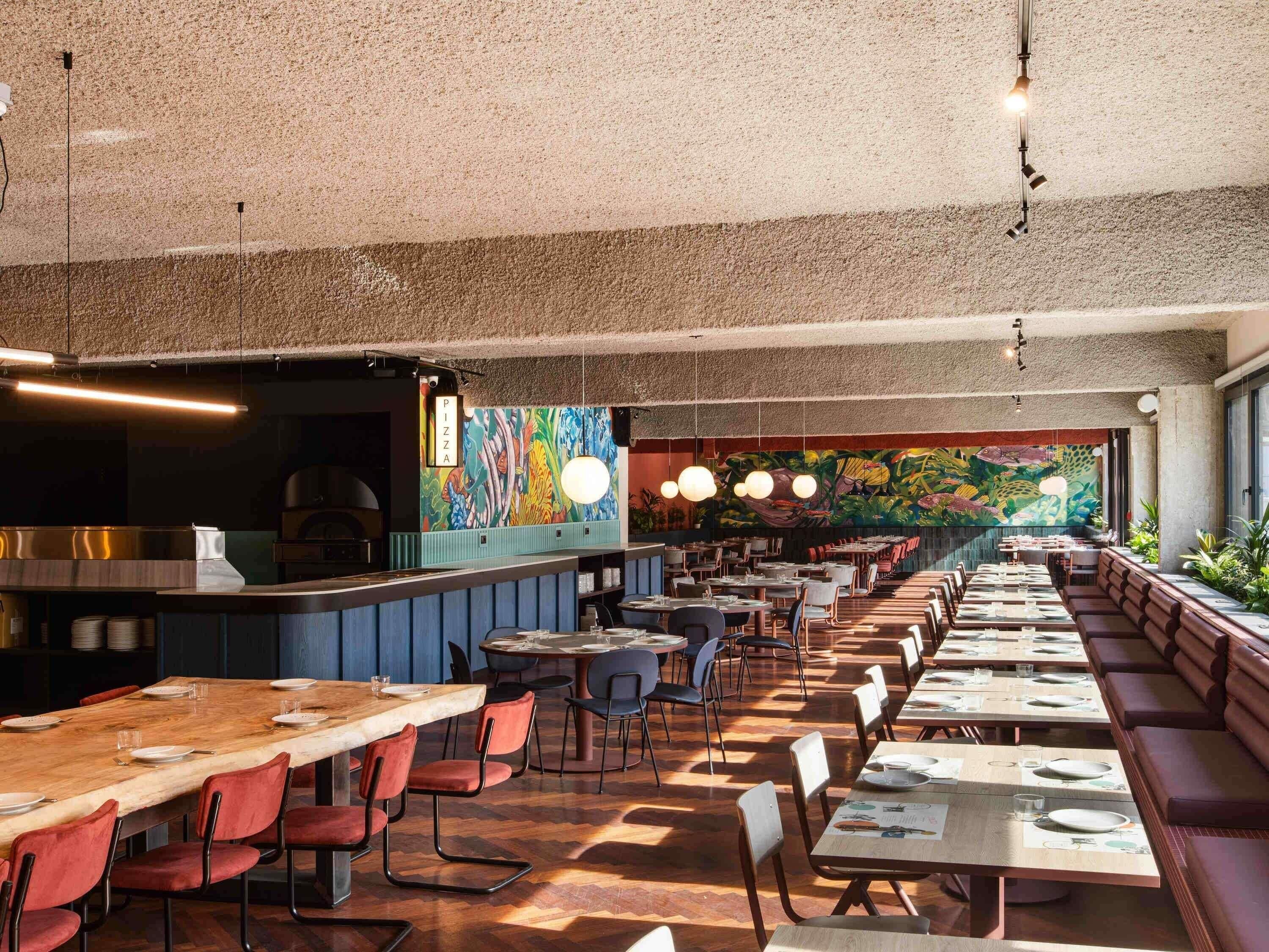 A restaurant interior with long communal tables, booths, and a colorful mural on the back wall.
