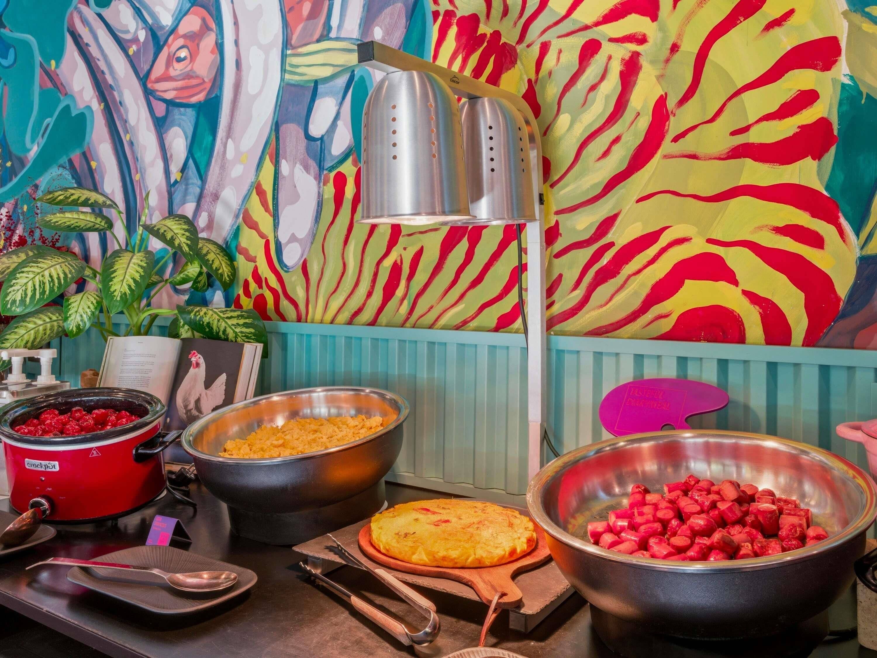 Een keukentafel met een rode slowcooker, een grote metalen kom, een houten snijplank met een gebakken gerecht en een schaal met in blokjes gesneden rode groenten.