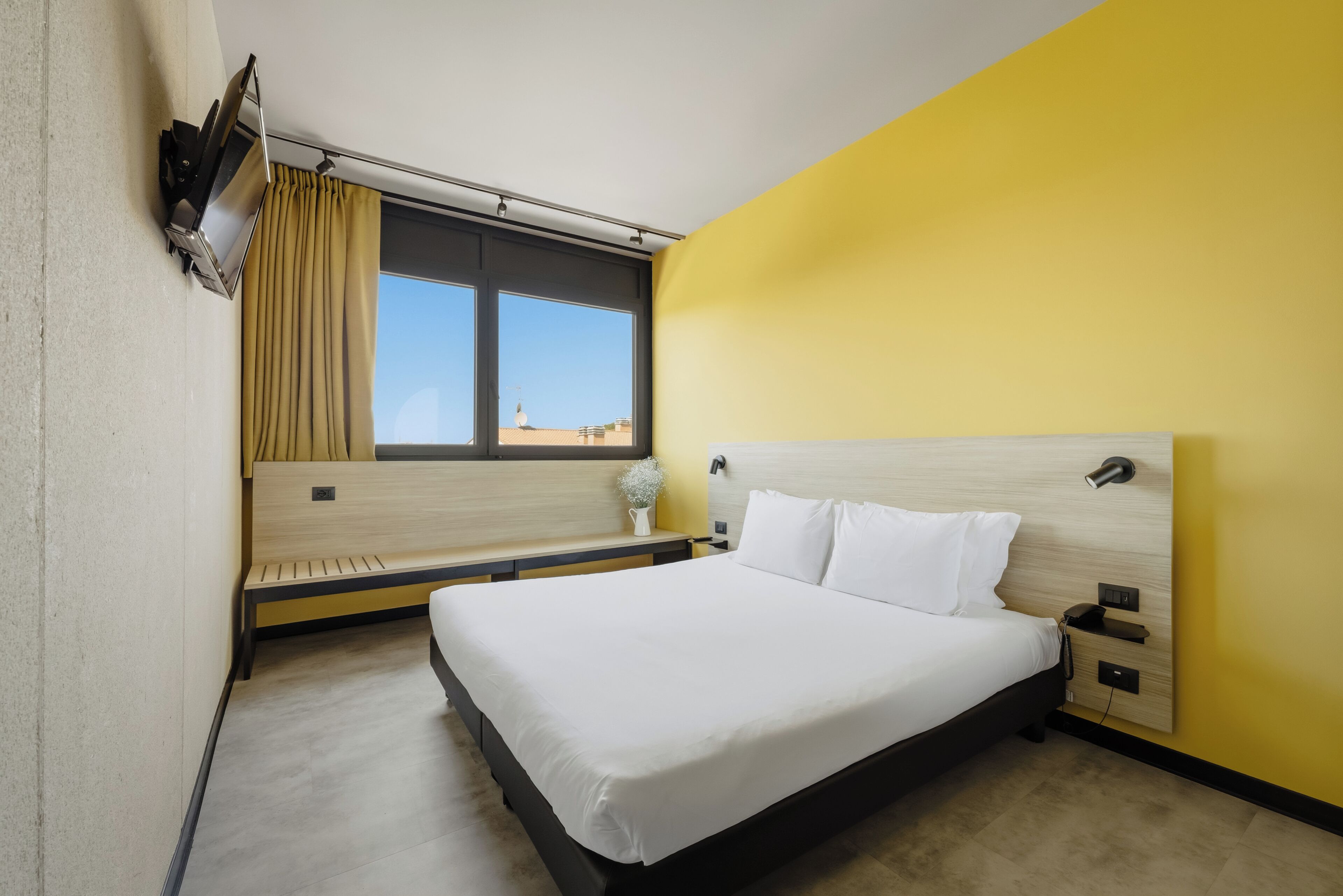 A hotel room with a bed, bedside table, lamp, window with curtains, and a view of the cityscape.
