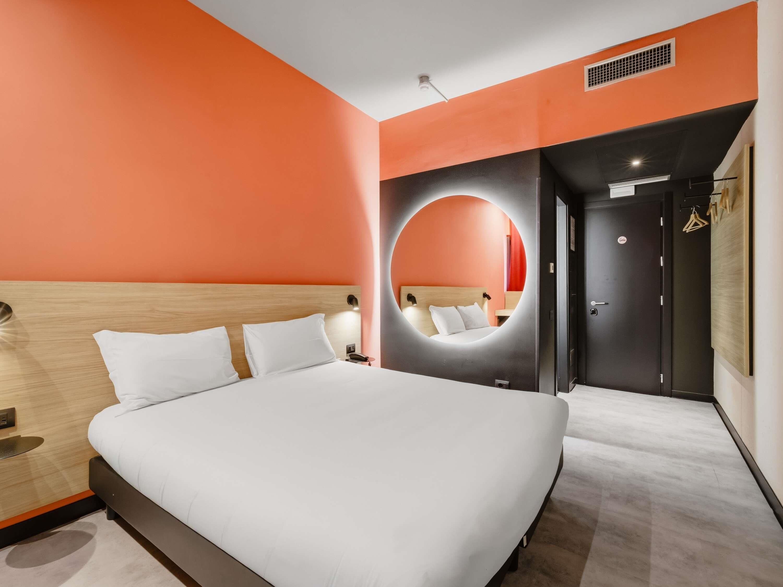 A modern hotel room with a large bed, a round mirror, and a door with a coat rack.