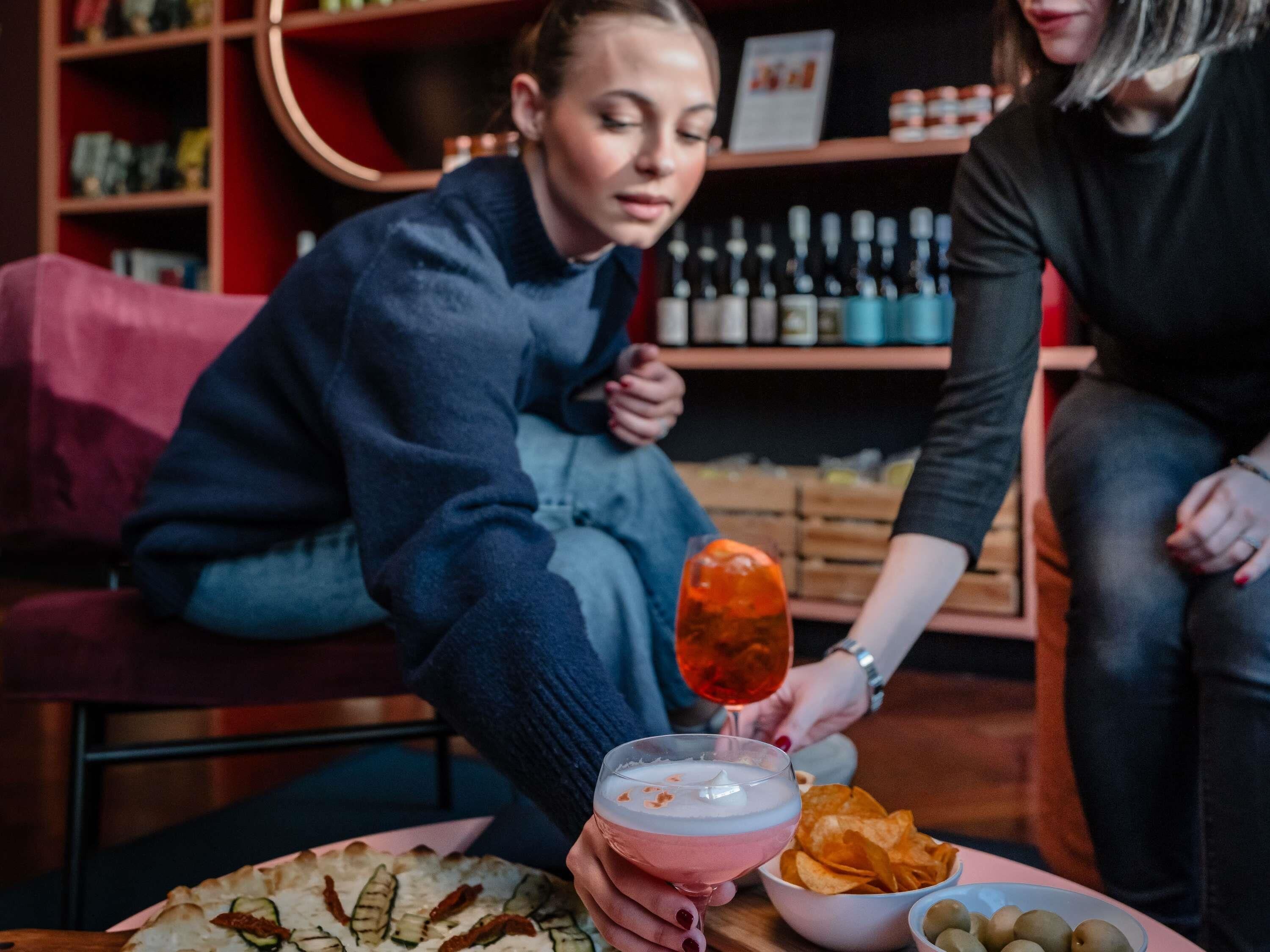 A person holding a drink, seated at a table with a plate of food and a bowl of chips.