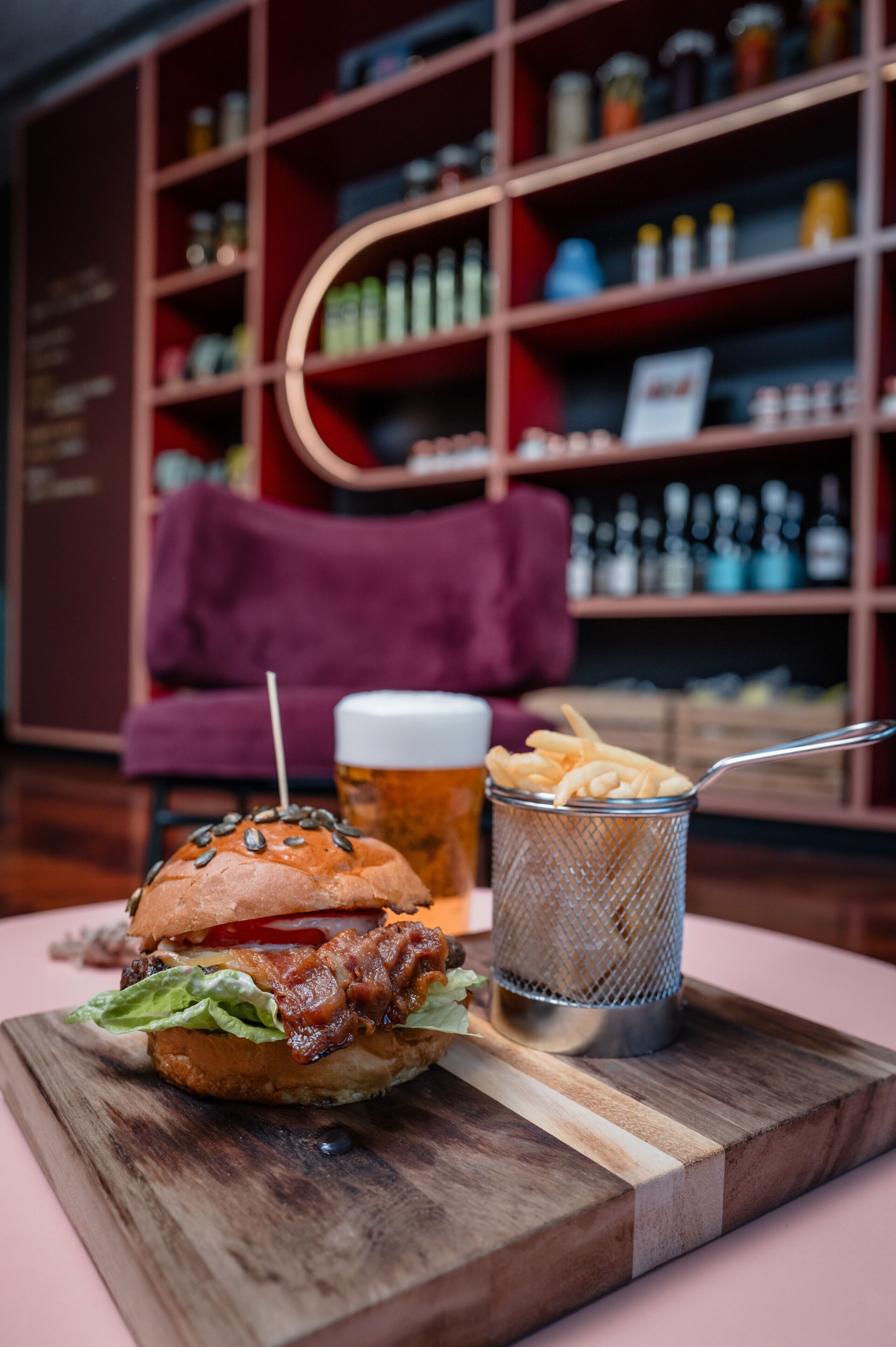 A burger with fries on a wooden board, a glass of beer, and a metal strainer with fries in the foreground.