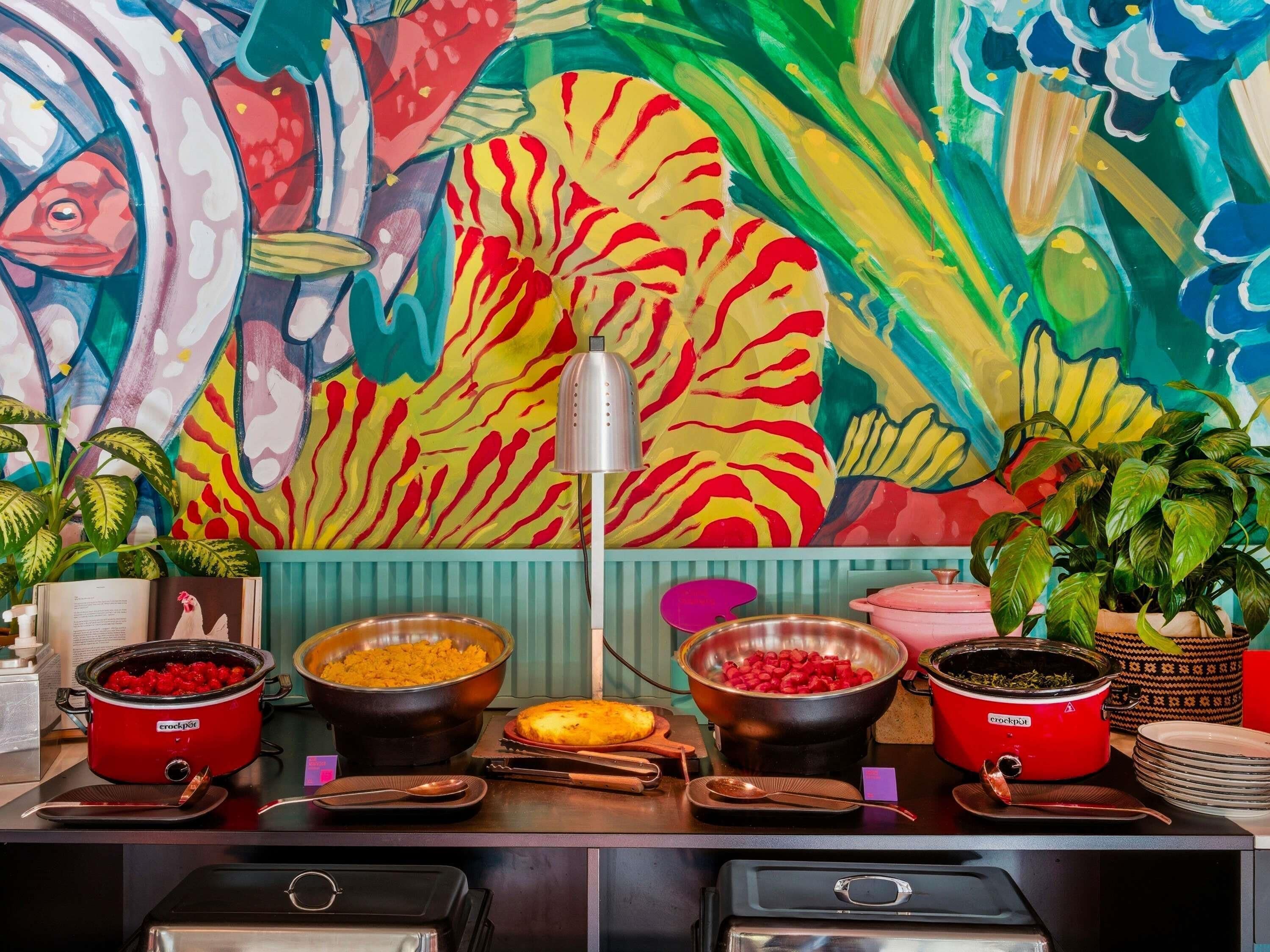 A buffet setup with various food items, a crockpot, and a colorful mural in the background.