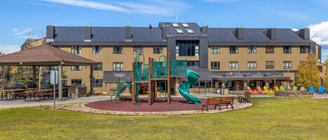 A playground with slides and climbing structures, surrounded by a building with multiple windows and a flat roof.